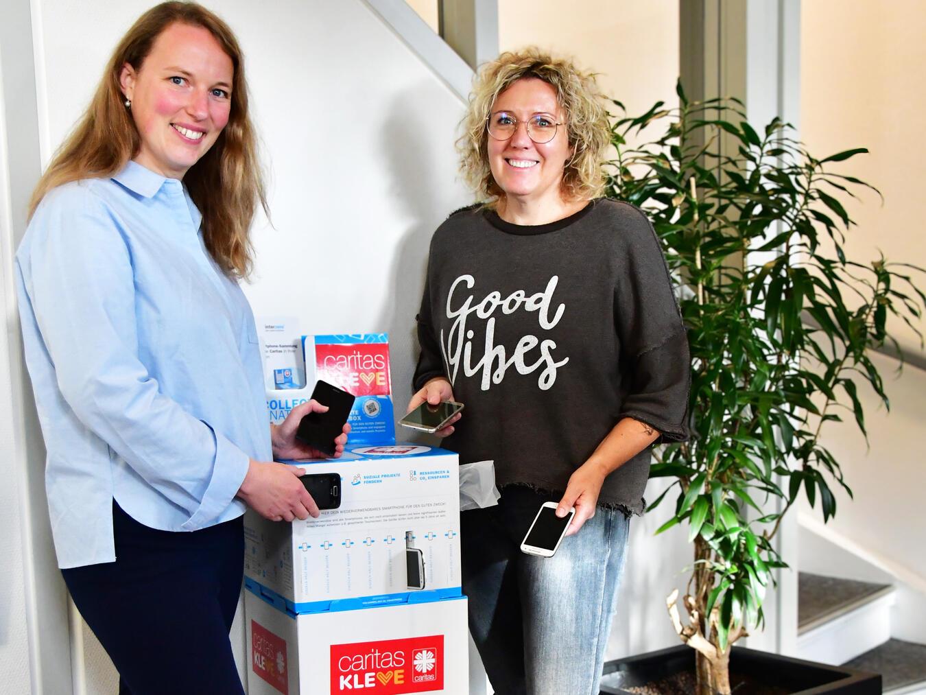 Anne-Kathrin Lehmann, Fachdienstleiterin Gemeindecaritas & Ehrenamtskoordination, und Nina Narzynski vom Fachbereich Soziale Hilfen präsentieren die Handysammel-Box im Caritas-Beratungszentrum in Kleve. Foto: Caritas