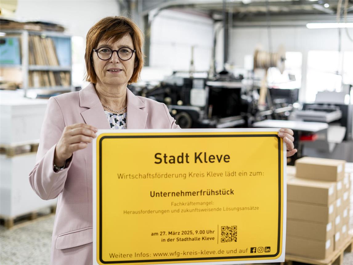 Brigitte Jansen, Geschäftsführerin der Wirtschaftsförderung Kreis Kleve, präsentiert das Ortsschild der Stadt Kleve mit der Einladung zum Unternehmerfrühstück. Foto: WfG Kreis Kleve