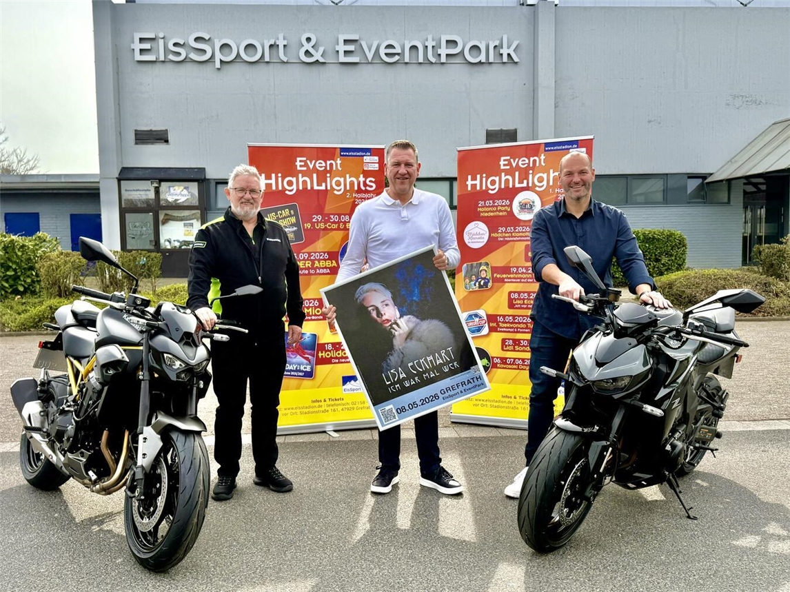 Drei Männer posieren mit zwei schwarzen Motorrädern vor zwei roten Aufstellern. Auf diesen ist das Eventsprogramm des Eisstadions Grefrath zu lesen. 