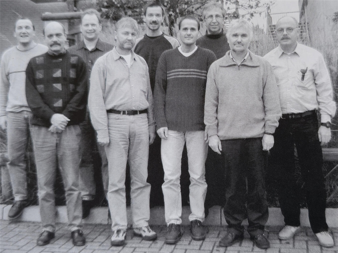 "Gruppenfoto: Team der ersten Stunde im Kreis Kleve, acht Männer in zwei Reihen, lächelnd, Geschäftskleidung"