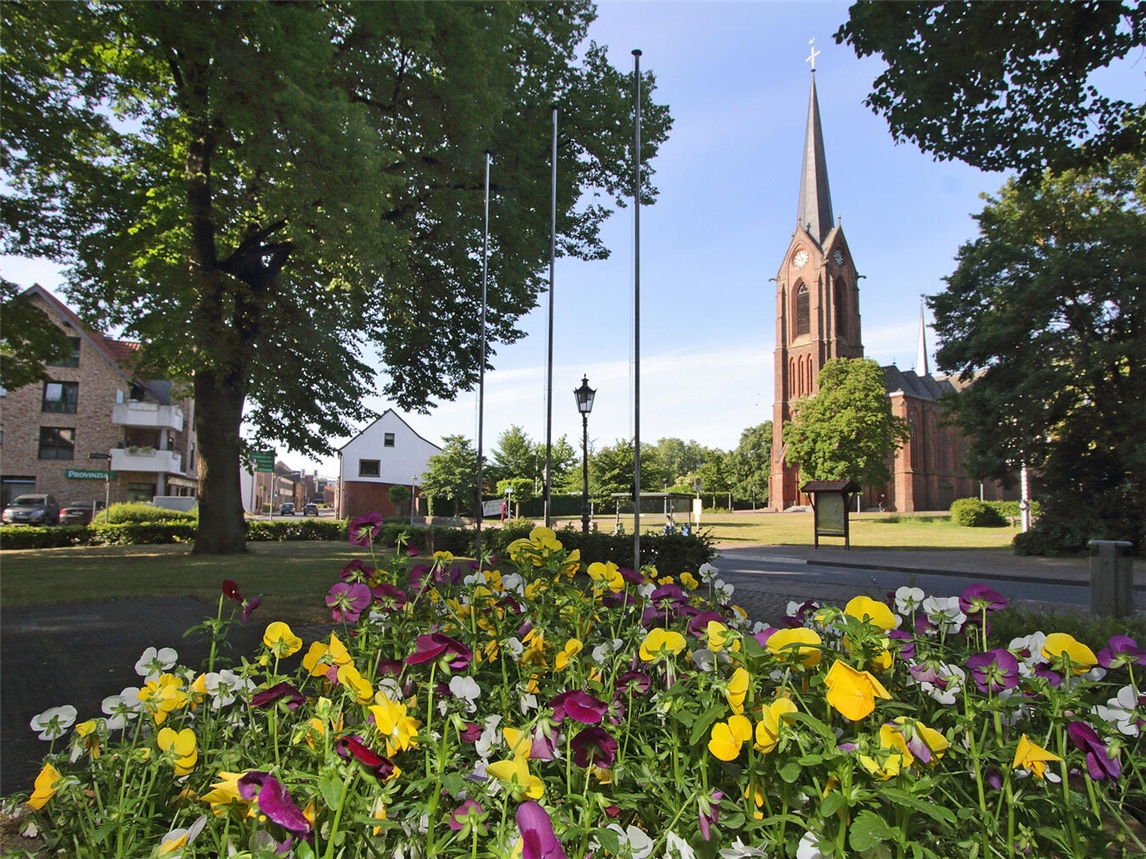 St. Hubertus-Kirche in Rheurdt-Schaephuysen. Foto zeigt einen grünen Park mit vielen lila und gelben Blumen im Vordergrund. Im Hintergrund ist die Kirche mit hohem, spitzen Kirchturm.