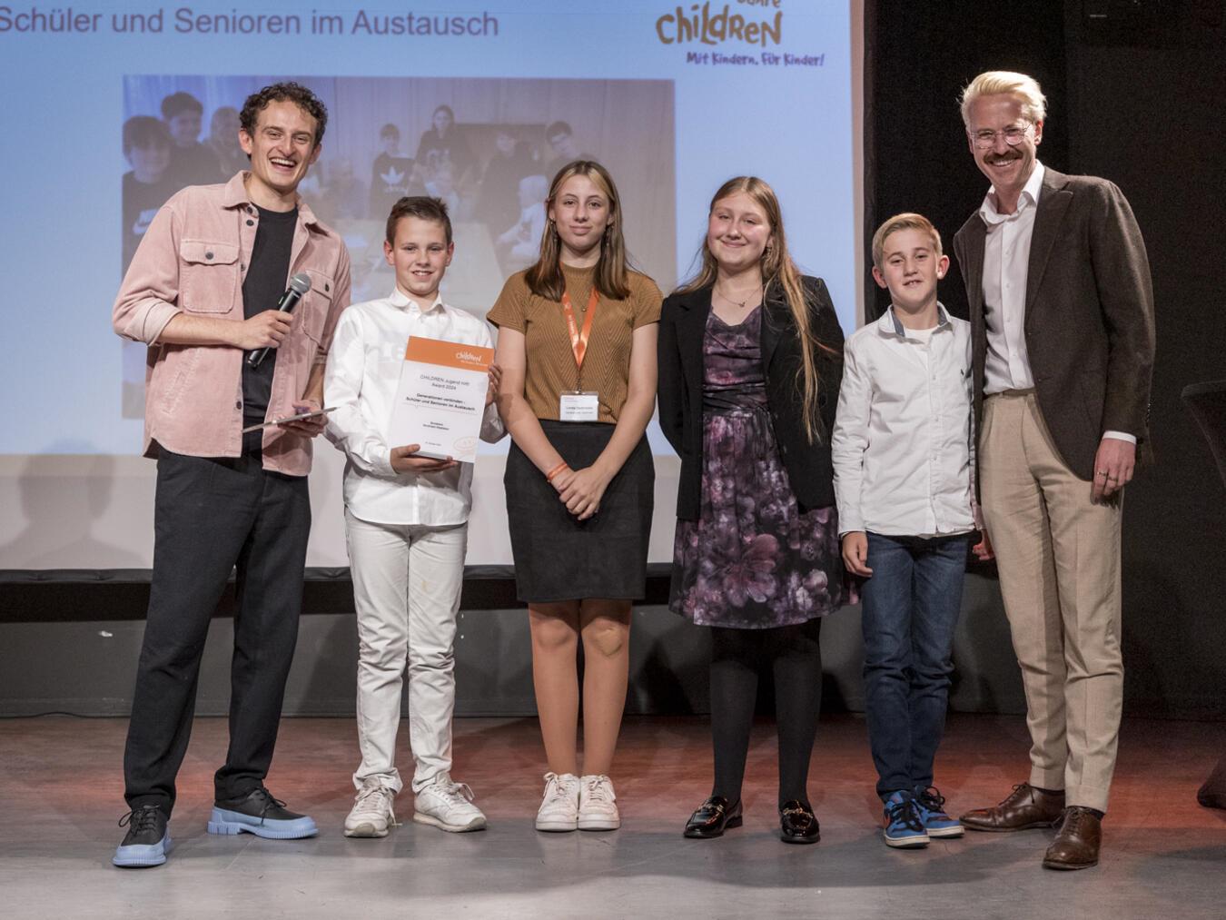 Die Projektteilnehmenden aus Sonsbeck mit Moderator Julian Jannsen („Checker Julian“, l.) und dem „Children for a better World“-Vorstandsvorsitzendem Raphael Langenscheidt (r.). Foto: Merlin Nadj-Torma