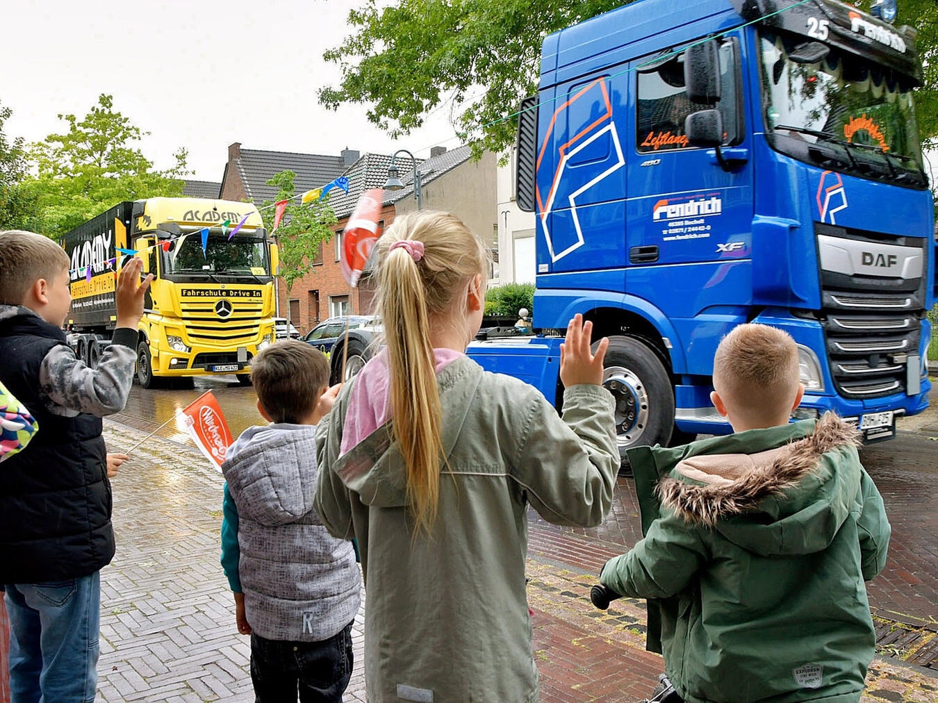 Menschen jubeln Lkw-Kolonne beim WOW-Run zu, Fokus auf kleine Beifahrer, fröhliche Atmosphäre, Straßenfest, Gemeinschaftsveranstaltung.