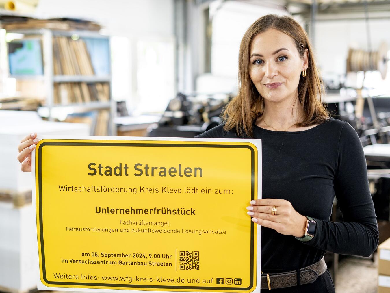 Fee Knupp von der Wirtschaftsförderung Kreis Kleve präsentiert das Ortsschild der Stadt Straelen mit der Einladung zum Unternehmerfrühstück.Foto: WfG Kleve