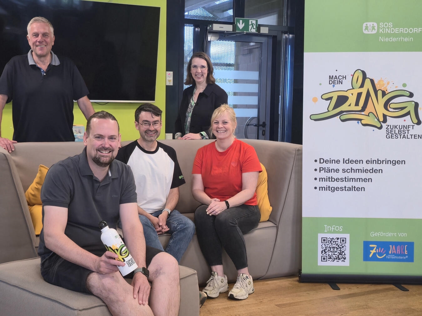 Team des Projekts „Mach dein Ding“ lächelt für ein Gruppenfoto. Im Vordergrund Ideenbegleiter, im Hintergrund Leitung von SOS-Kinderdorf.