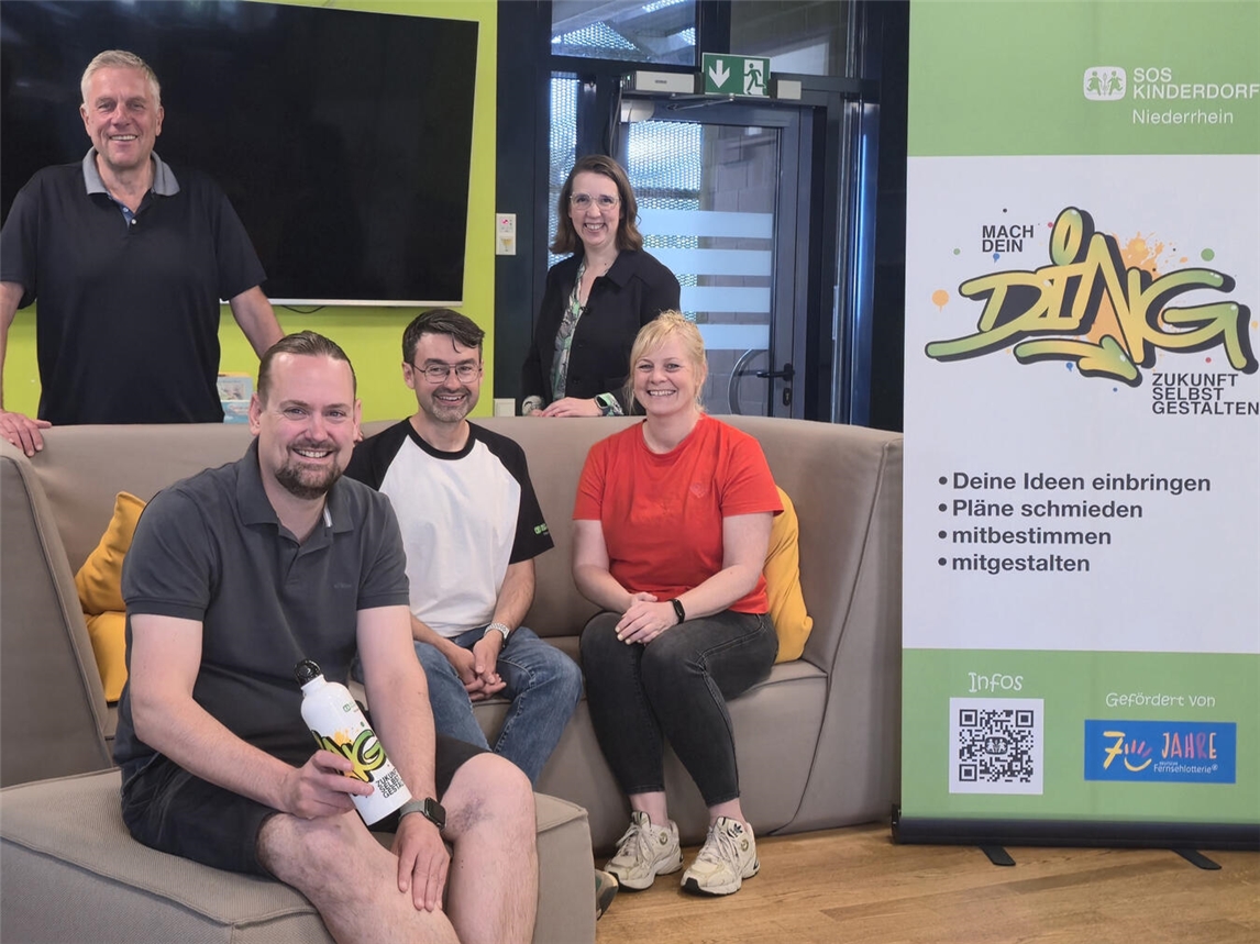 Team des Projekts „Mach dein Ding“ lächelt für ein Gruppenfoto. Im Vordergrund Ideenbegleiter, im Hintergrund Leitung von SOS-Kinderdorf.