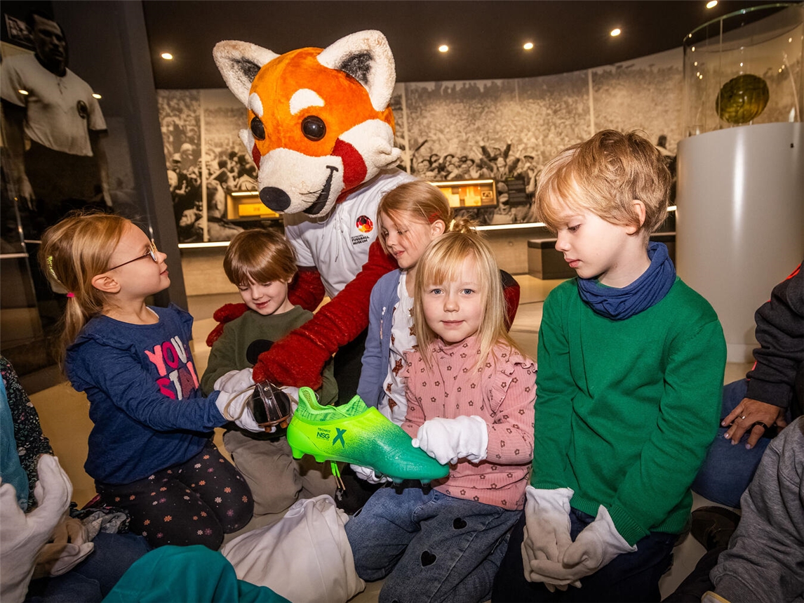 Fußballgeschichte zum Anfassen: Maskottchen Rio zeigt Kindern Fußballschuhe aus verschiedenen Jahrhunderten.