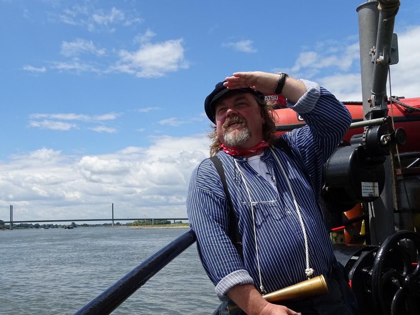 "Heinz Wellmann als ‚Hein vom Rhein‘ in traditioneller Kleidung in der Stadt Re, lächelnd, bei sonnigem Wetter."