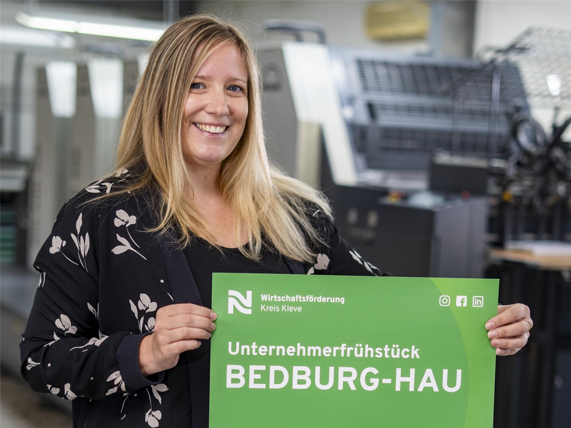 Irina Janßen von der Wirtschaftsförderung Kreis Kleve präsentiert das Plakat mit der Einladung zum Unternehmerfrühstück in Bedburg-Hau. Foto: WFG/Gerhard Seybert