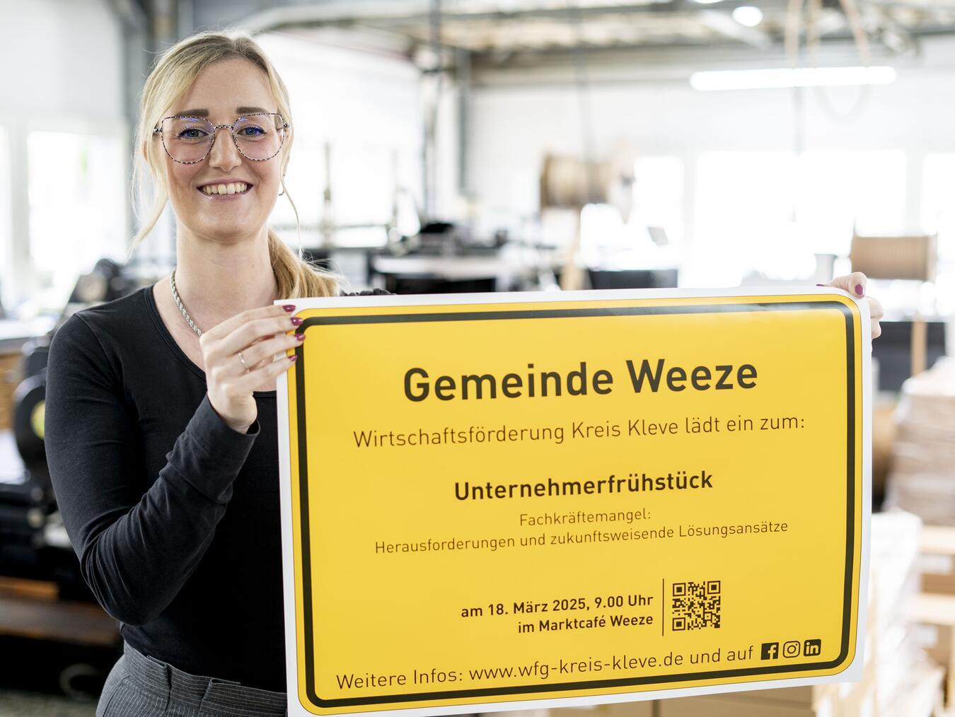 Irina Schultz von der WFG Kreis Kleve präsentiert das Ortsschild der Gemeinde Weeze mit der Einladung zum Unternehmerfrühstück.