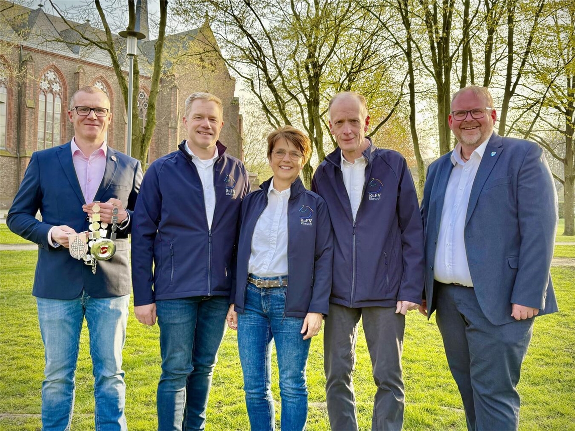 Die Festkettenträgerin steht mittig zwischen ihren beiden Adjutanten. Alle tragen Jeans, weiße T-shirts sowie ihre dunkelblaue Vereinsjacke mit dem RuF-Pferdelogo. Links neben ihnen steht der Vorsitzende der Veeinsgemeinschaft, er hält die Festkette. Rechts steht Bürgermeister Dirk Katelaers. Alle stehen im Grünen vor einer Kirche. 