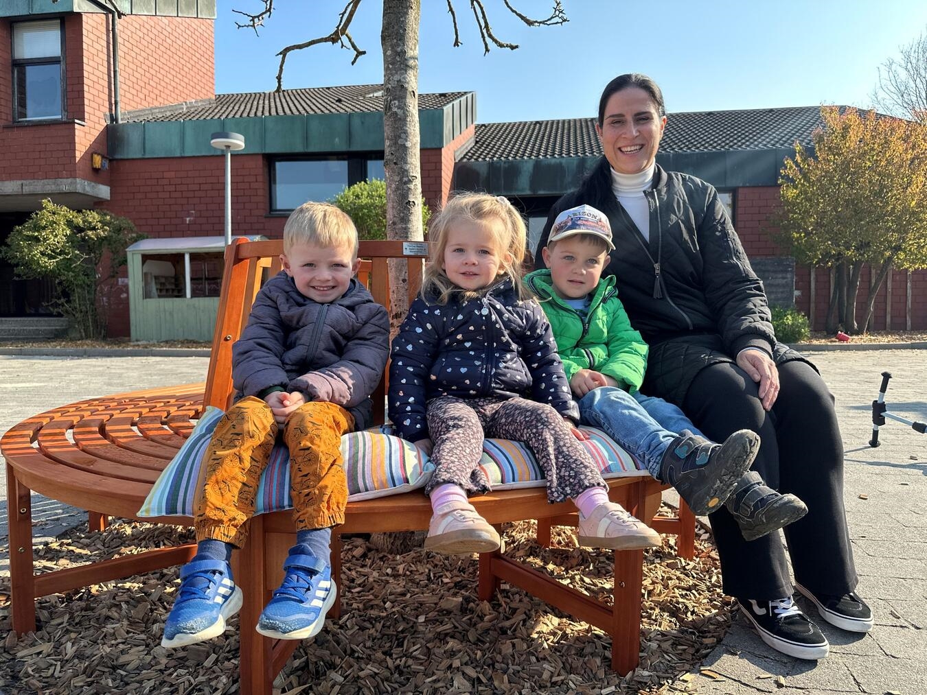 "Kita-Leiterin Martina van Offern mit Kindern auf neuer Holzbank in Kita am Wald, glückliche Gesichter, Gemeinschaftsprojekt"