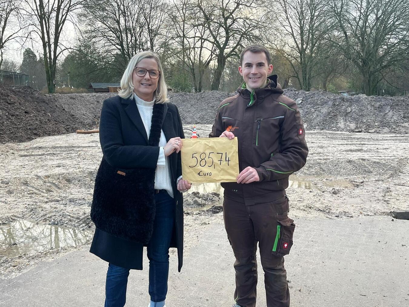 Bedburg-Hau: OGS-Kinder spenden für den Klever Tiergarten Manuela Witzke übergibt die Einnahmen des Bücherflohmarktes an Martin Polotzek. Das Geld fließt in den Umbau der Trampeltieranlage, die im Hintergrund zu sehen ist. Foto: CV Kleve