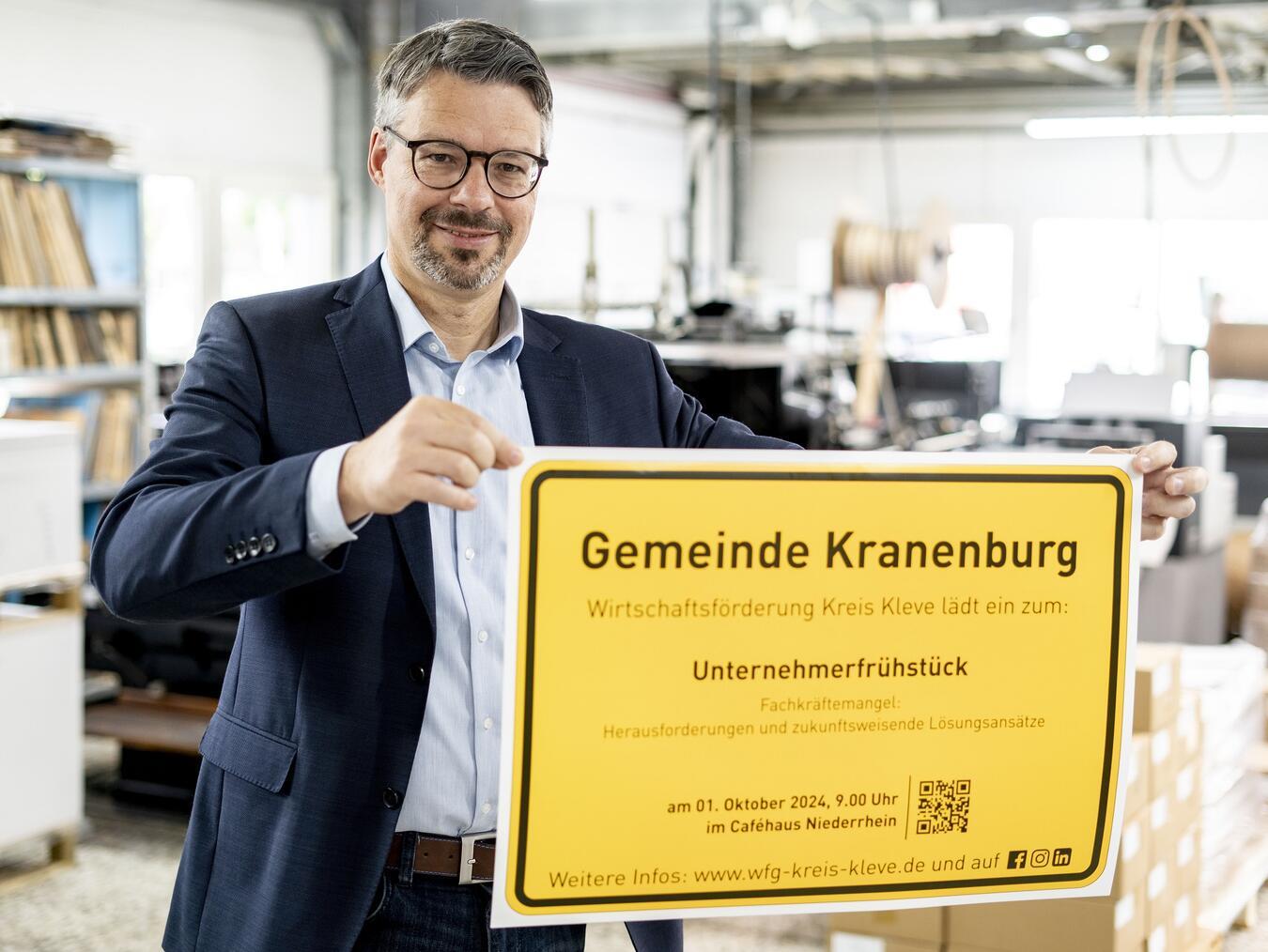 Marc Cattelaens von der Wirtschaftsförderung Kreis Kleve präsentiert das Ortsschild der Gemeinde Kranenburg mit der Einladung zum Unternehmerfrühstück. Foto: Wirtschaftsförderung