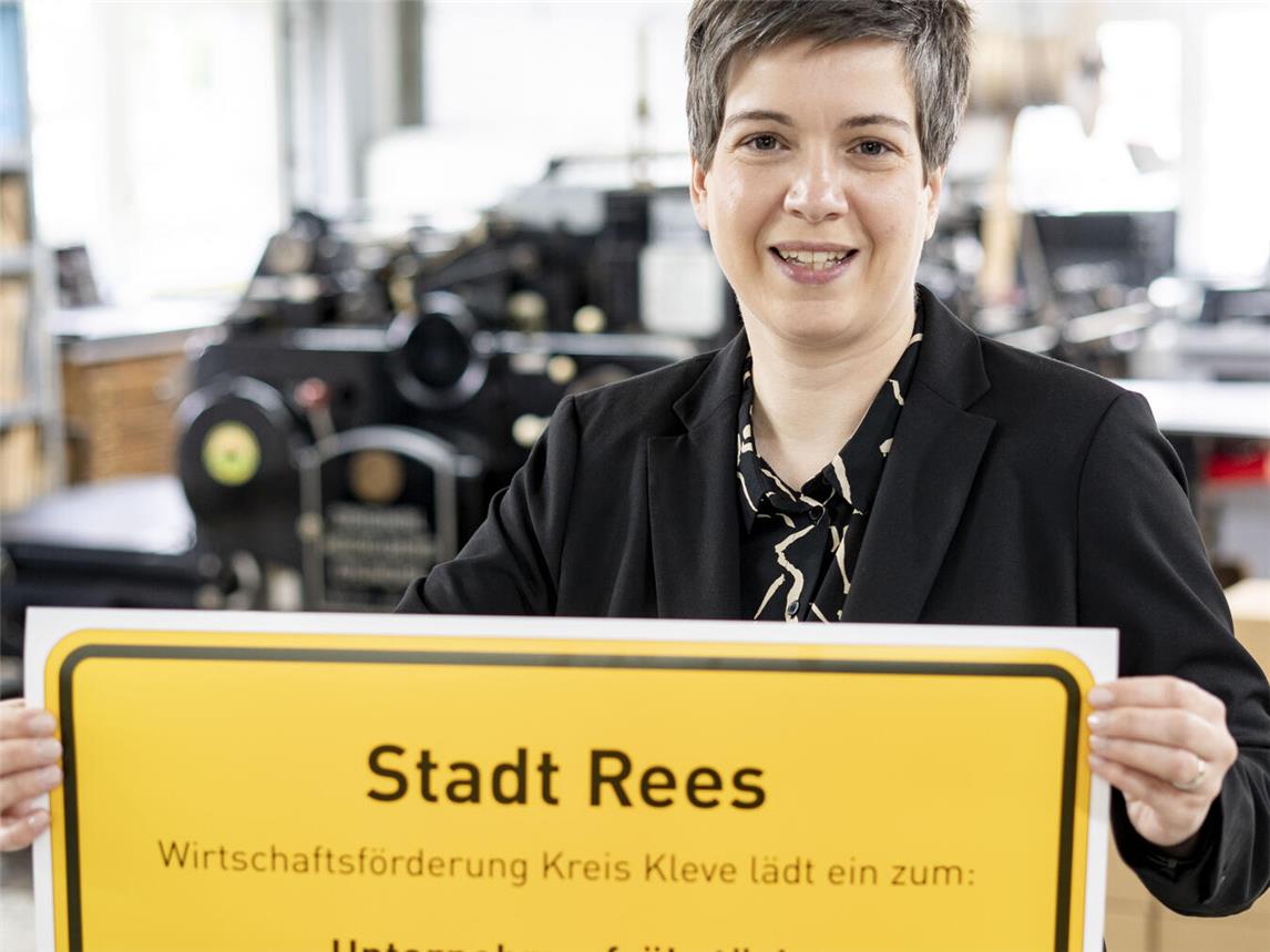 Nathalie Tekath-Kochs von der Wirtschaftsförderung Kreis Kleve präsentiert das Ortsschild der Stadt Rees mit der Einladung zum Unternehmerfrühstück. Foto: WFG Kreis Kleve