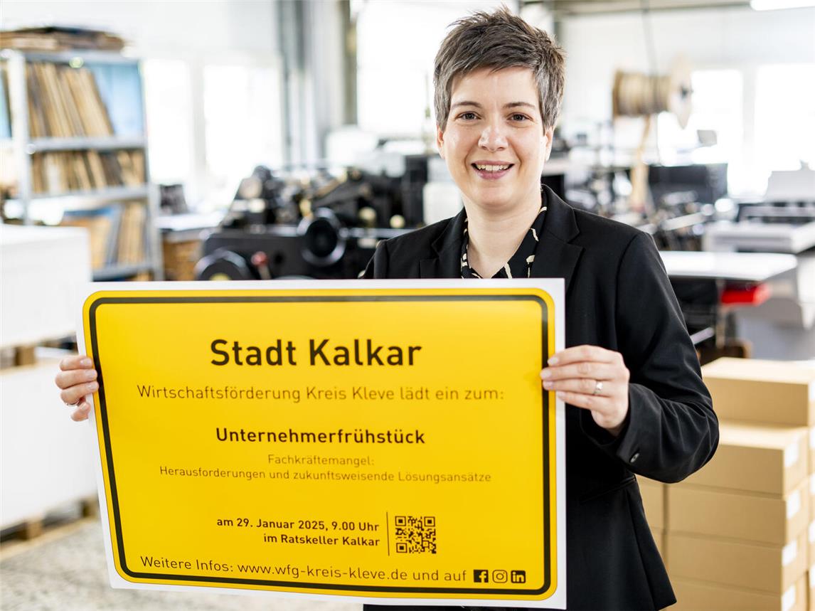 Nathalie Tekath-Kochs von der Wirtschaftsförderung Kreis Kleve präsentiert das Ortsschild der Stadt Kalkar mit der Einladung zum Unternehmerfrühstück. Foto: WfG / Gerhard Seybert