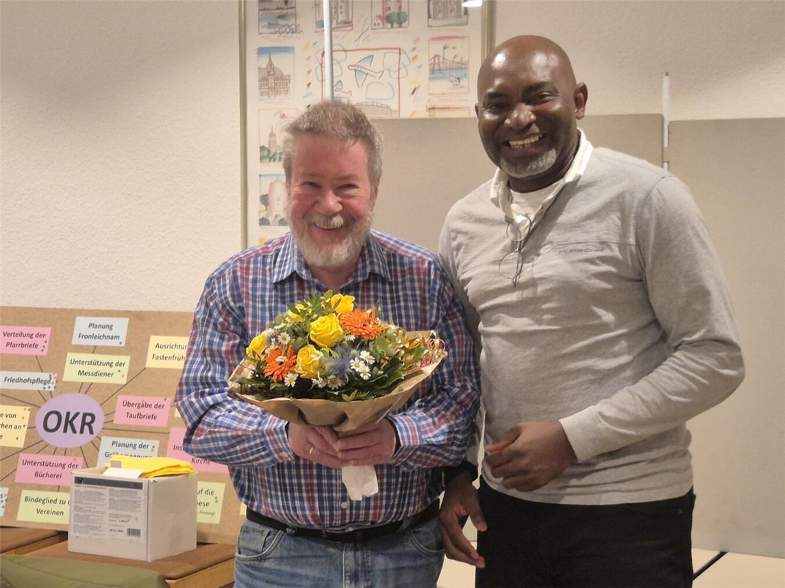 Sie lächeln von ganzem Herzen, wenngleich dieser Moment für beide mit Wehmut verbunden ist: Theo Kersten (l.) erhält von Pfarrer „Uche“ Aba Blumen und aufrichtige Dankesworte als Anerkennung für seine langjährige Arbeit. Foto: Gudrun Beaupoil (OKR)