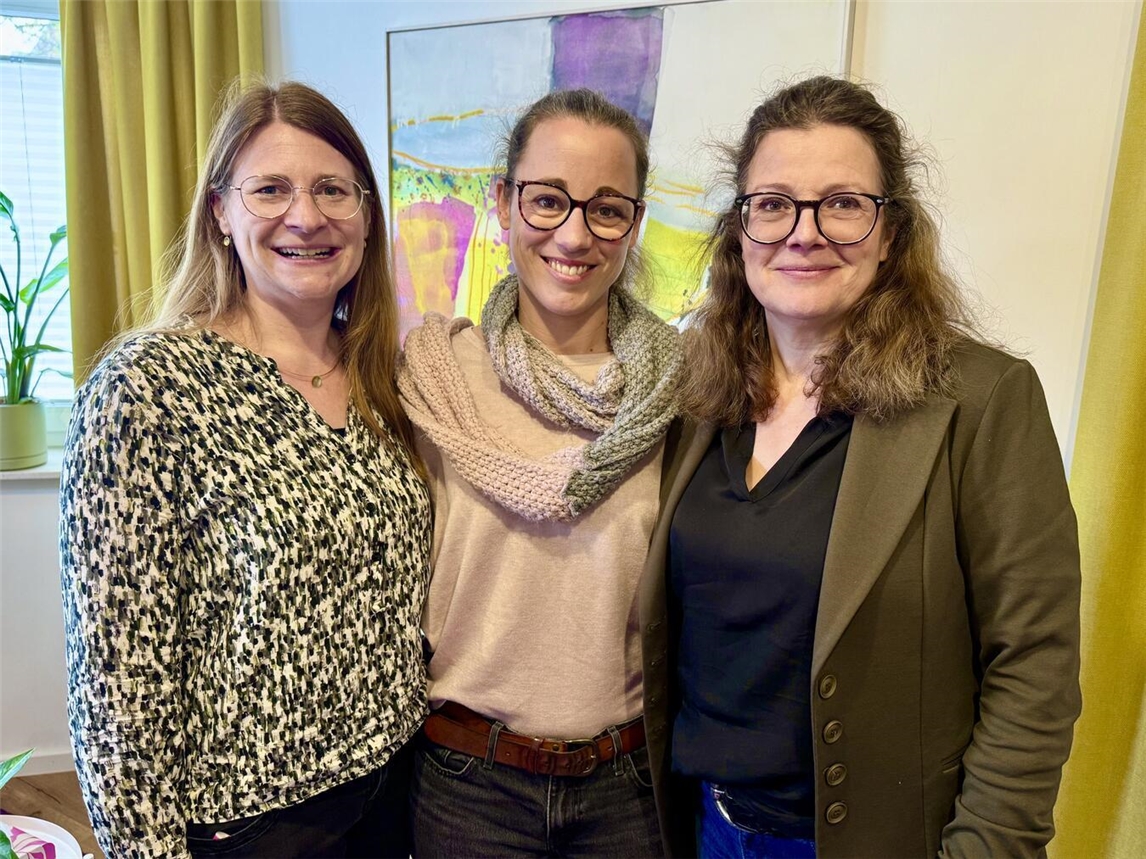 Drei Frauen stehen Arm in Arm vor einem Gemälde in den Farben lila, gelb und pink. Rechts und links befinden sich gelbe Vorhänge. 