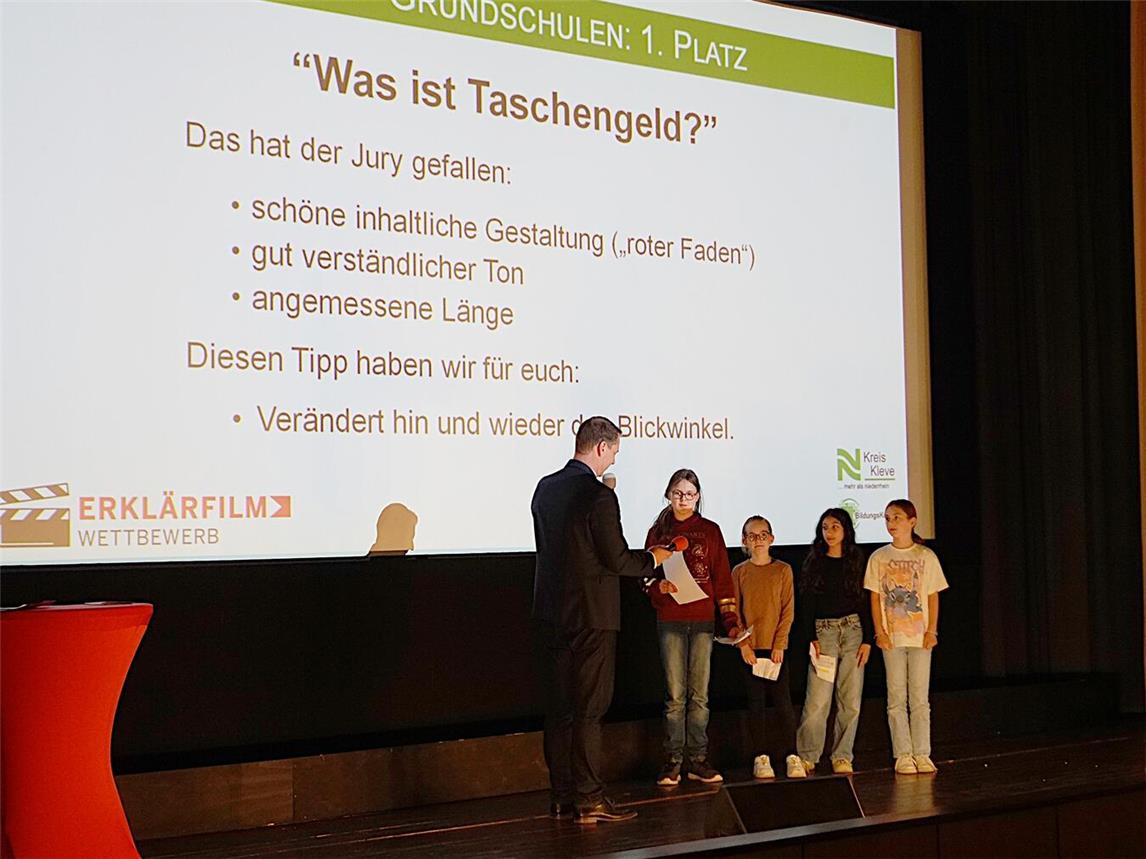 „Was ist Taschengeld?“ – das erklärt die Schülergruppe der Leegmeer-Grundschule Emmerich am Rhein in ihrem Kurzfilm. Schulrat Andreas Czymay überreicht den Mädchen und Jungen den ersten Preis des Erklärfilm-Wettbewerbs Kreis Kleve. Foto: Kreis Kleve